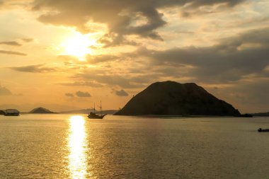 Golden hour in Komodo National Park, Indonesia. Sun sets behind one of the islands, leaving a golden path on the calm surface of sea. Some boats anchored to the shore, enjoying the natural spectacle.