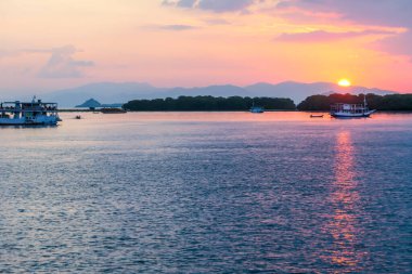 Golden hour in Komodo National Park, Indonesia. Sun sets behind one of the islands, leaving a golden path on the calm surface of sea. Some boats anchored to the shore, enjoying the natural spectacle.