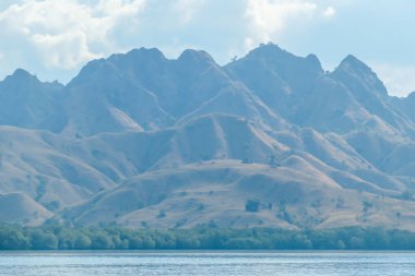 A view on idyllic Komodo Island, Indonesia. Volcanic island is barely overgrown with plants. There are few clouds above the island. Calm and clear surface of the sea. Perfect day for sailing