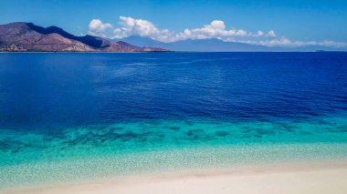 A drone shot of a small island near Maumere, Indonesia. The coastal line changes colors from white to turquoise and navy blue. There are some islands in the back. Hidden gem of Indonesia. Paradise