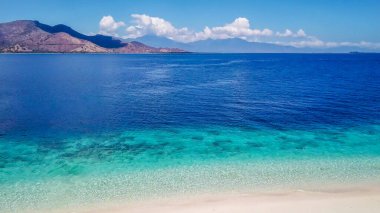 A drone shot of a small island near Maumere, Indonesia. The coastal line changes colors from white to turquoise and navy blue. There are some islands in the back. Hidden gem of Indonesia. Paradise