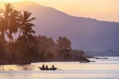 A sunset in Maumere, Flores, Indonesia. The sun sets behind the slope of the island. Serenity and calmness. There are few fishermen fishing from their boats in the bay. Palm trees growing on the shore