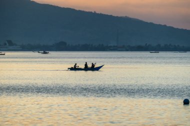 A sunset in Maumere, Flores, Indonesia. The sun sets behind the slope of the island. Serenity and calmness. There are few fishermen fishing from their boats in the bay. Palm trees growing on the shore