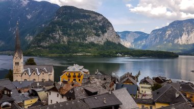 A small village located by the lake in Hallstatt, Austria. There is a tall church tower in the middle of the village. Alpine village. Idyllic landscape. High mountains rising from the lake.