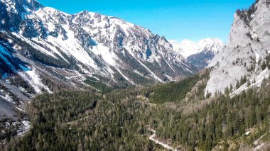 A drone shot of an Alpine landscape in Austria. There are few mountain ranges surrounding the area. Bright and sunny day. Higher parts of the mountains covered with snow. Pathway through the forest