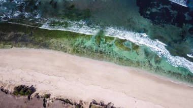 A done shot of Nyang Nyang Beach, Bali, Indonesia. The waves are rushing to the shore, making the water bubbly. The beach is covered with green algae, further on it's sandy. Idyllic hidden beach