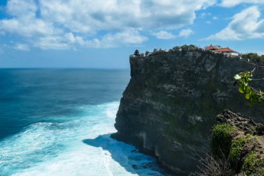 A view on Uluwatu Cliffs, Bali, Indonesia. There is a small temple on the top of the cliff. The waves are rushing to the shore, making the water bubbly. Steep and dangerous fall. Hidden gem