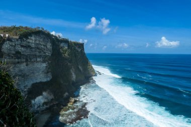A view on Uluwatu Cliffs, Bali, Indonesia. The waves are rushing to the shore, making the water bubbly. The cliffs are overgrown with small plants and moss. Steep and dangerous fall. Hidden gem
