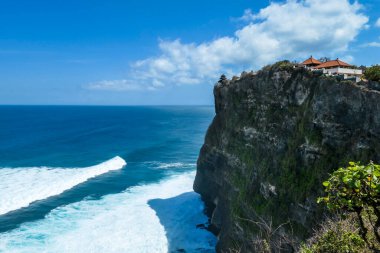 A view on Uluwatu Cliffs, Bali, Indonesia. There is a small temple on the top of the cliff. The waves are rushing to the shore, making the water bubbly. Steep and dangerous fall. Hidden gem