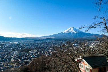 A distant view on Mt Fuji in Japan on a clear, wintery day. The top parts of the volcano are covered with a layer of snow. Holly mountain of Japan. A few tree branches and a house in the frame.