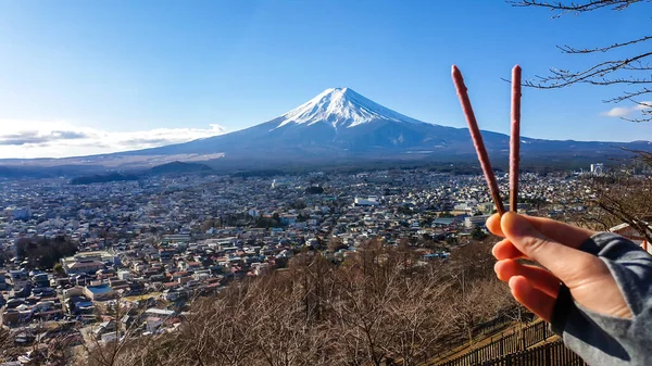 A hand holding two chocolate sticks, making a V-shape, with a distant view on Mt Fuji in Japan on a clear, wintery day. The top parts of the volcano are covered with a layer of snow. Holly mountain