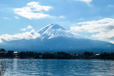 An idyllic view on Mt Fuji from the side of Kawaguchiko Lake, Japan. The mountain is surrounded by clouds. Serenity and calmness. The top of the volcano is covered with snow. Calm surface of the lake.