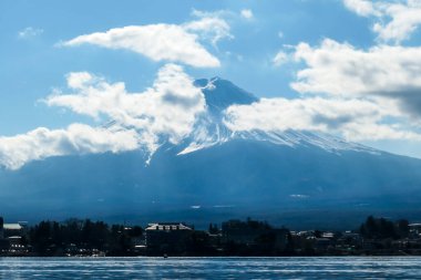 An idyllic view on Mt Fuji from the side of Kawaguchiko Lake, Japan. The mountain is surrounded by clouds. Serenity and calmness. The top of the volcano is covered with snow. Calm surface of the lake.