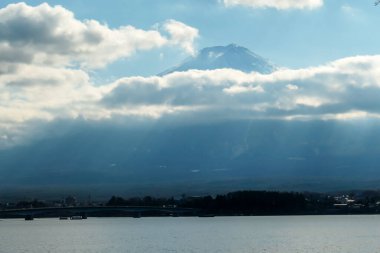 A close up view on Mt Fuji from the side of Kawaguchiko Lake, Japan. The mountain is hiding behind the clouds. Top of the volcano covered with a snow layer. Serenity and calmness. Calm lake's surface