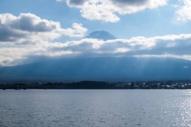 A close up view on Mt Fuji from the side of Kawaguchiko Lake, Japan. The mountain is hiding behind the clouds. Top of the volcano covered with a snow layer. Serenity and calmness. Calm lake's surface