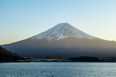 A close up view on Mt Fuji from the side of Kawaguchiko Lake, Japan. Soft colors of sunset - golden hour. Top of the volcano covered with a snow layer. Serenity and calmness. Calm lake's surface