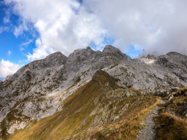 A pathway leading to high peaks in Italian Alps. Sharp slopes on both sides of the valley. Hard to reach mountain peaks. There are many mountain ranges in the back. Serenity and peace. Autumn vibes