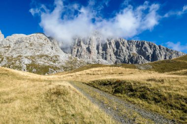 A pathway leading to high peaks in Italian Alps. Sharp slopes on both sides of the valley. Hard to reach mountain peaks. There are many mountain ranges in the back. Serenity and peace. Autumn vibes