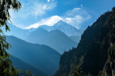 View on Himalayan valley along Annapurna Circuit Trek, Nepal. There is a dense forest in front. High, snow caped mountains' peaks catching the sunbeams. Serenity and calmness. Barren slopes