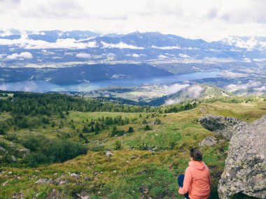 Girl squating on the ground, admiring the view on the MIllstaeter lake in Millstatt, Granattor, Austria. Clouds in the valley. Dense forest around. Serene landscape. HIgh mountains. Alpine meadows