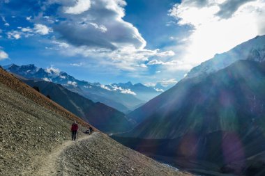 Girl wearing sunglasses, pink jacked and black leggins, walks on the trail in  Annapurna Circuit Trek, Himalayas, Nepal. Few trekkers behind her. Endless chains of mountains. Rocky trail.
