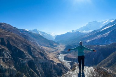 Girl wearing a beanie and blue jumper, spreads her arms wide, breathing deeply the fresh mountain air. Her gesture represents freedom and happiness. Below a long valley stretches in Himalayas.