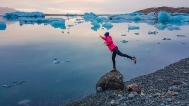 A girl wearing pink jacket stands at the rock by the glacier lagoon, forms a flying superhero pose. Empowered woman. Blue icebergs slowly drifting towards the sea. Effect of global warming.