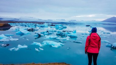 A young woman standing on the side of the glacier lagoon. Huge and massive ice bergs are slowly drifting towards the sea. Girl wears pink jacket. Pink contrasted with cold blue tones. Global warming