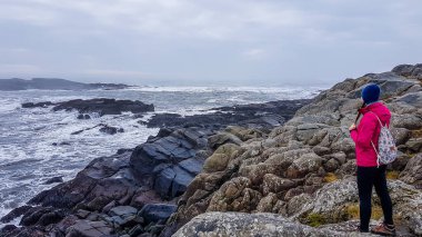 A girl wearing pink jacket and a backpack, standing at the stony shore and looking at the sea. Rough surface of the sea, waves struggle through the rocks to get to the shore. Solo traveling girl.