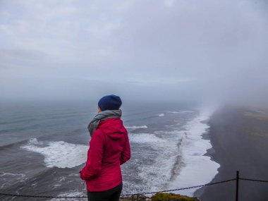 A girl wearing a pink jacket and a beanie standing at the top of the cliff with her hands in the pockets, looking at the rough sea below. Waves wash the black sand beach abruptly. Moody and mysterious