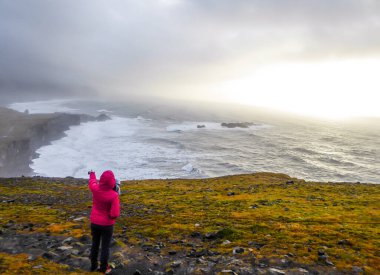 A girl wearing a pink jacket standing at the top of the cliff, pointing at the rough sea in front of her. Waves wash the black sand beach abruptly. Moody and mysterious atmosphere. Sun slowly setting