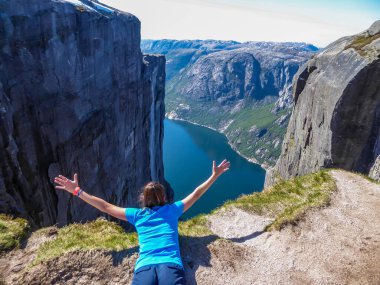A girl lying at the edge of a steep mountain pretends to be flying. She spreads arms wide, in a gesture of freedom. In front of her stunning Lysefjorden shimmering with many shades of blue and green