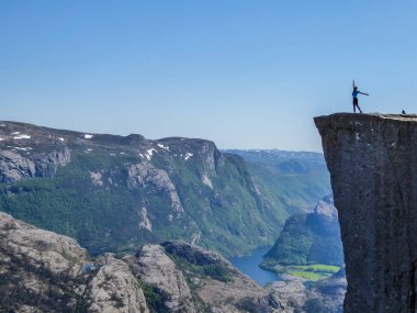 A girl wearing blue shirt stands at the edge of a steep cliff of Preikestolen, Pulpit Rock. A view on Lysefjorden. Girl enjoys the view, feeling free and happy, she spreads her arms wide open.