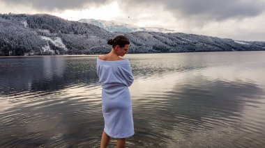 A girl in a white bathrobe walking in a shallow water of a lake. Half of the back of a girl is naked. On the other side of the lake there are mountains covered with snow. Winter alpine spa.