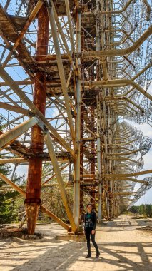 Girl standing under the Secret Soviet Radar 'DUGA-1', called a woodpecker hidden in the forest in the nearby of Prypiat city. Massive metal construction spreading on a wast territory.