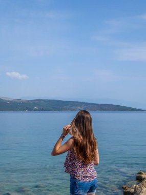 Girl with long hair stands at the shore of the Mediterranean Sea and looks at the calm surface of the water. She is calms and peaceful. Water has many shades of blue. In the back there are islands