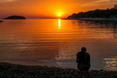 A girl enjoying the sunset by a beach. The sun sets over the horizon. The sun beams reflecting in the calm sea waters. There is an island on the side. The sky turns yellow. Girl enjoys the spectacle