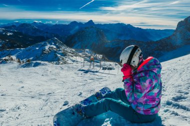 A snowboarder taking a break on the slope. The snowboard is white and blue, harmoniously fitting to the nature conditions - white snow and blue sky. There are endless chains of mountains in the back