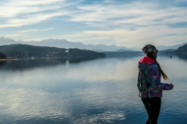 Girl wearing a jacket and beanie stands at the end of a promenade on the lake. Soft reflections of the clouds in the lake. Clear but cloudy day. High mountains in the back Calmness and relaxation