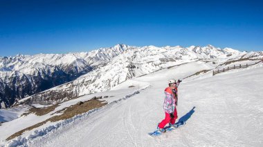 A snowboarder going down the slope in Heiligenblut, Austria. Perfectly groomed slopes. High mountains surrounding the girl, wearing pink trousers and colorful jacket. Girl wears helm for protection