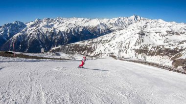 A snowboarder going down the slope in Heiligenblut, Austria. Perfectly groomed slopes. High mountains surrounding the girl, wearing pink trousers and colorful jacket. Girl wears helm for protection