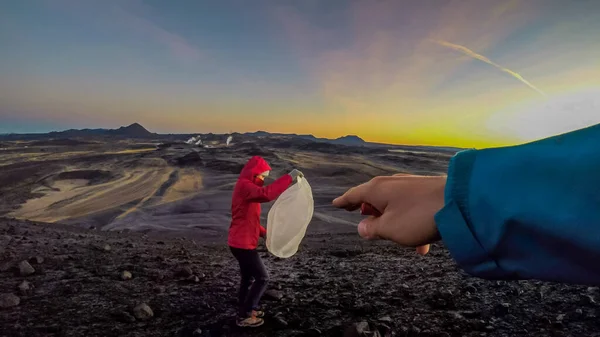 A hand pointing at a girl wearing pink jacket and holding a wish lantern. In the back sky is exploding with colors of sunrise. Harsh and barren landscape. Geothermal region, steam coming out of earth.