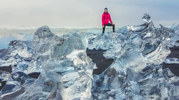 A close up on an ice structure, laying on the black sand beach in Iceland, diamond beach. A hazy silhouette of a girl wearing a pink jacket in the back. Sea throws the ice bergs on the shore.