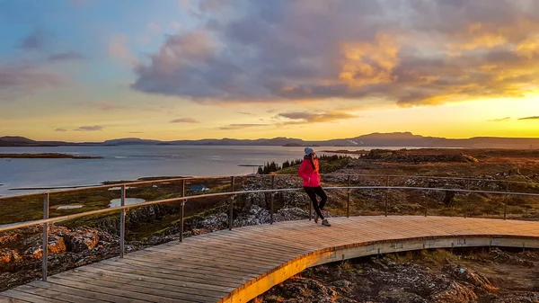 A beautiful girl wearing a pink jacket, leaning on a barrier during the sunset over national park Pingvellir in Iceland. Whole valley covered with golden hour\'s colors. Colors of explosion on the sky.