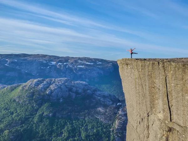 A girl wearing orange hoodie stands at the edge of a steep cliff of Preikestolen, Pulpit Rock. A view on Lysefjorden. Girl enjoys the view, feeling free and happy, she spreads her arms wide open.