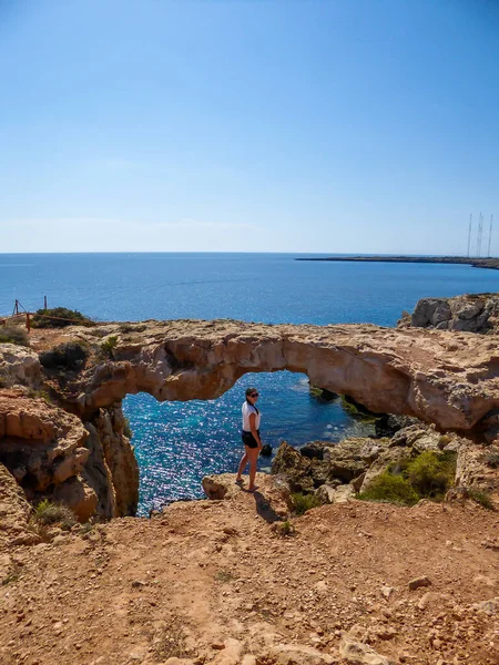 在塞浦路斯格拉科角陡峭的悬崖前 一个女孩站在一座石拱桥前 这座桥连接着两座悬崖 石层的巴伦斜坡 蓝色的海无边的地平线 — 图库照片