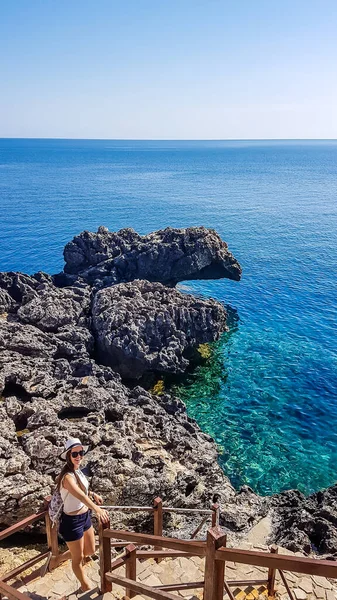 Girl wearing a hat, going down the stairs to a hidden beach, the staircase is surrounded by steep and sharp cliffs. Water shimmers with many shades of blue. Solo traveling adventurous girl.