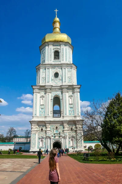 A young woman standing in front of St Sophia\'s Cathedral, Kiev, Ukraine. Golden rooftop of the tower. Play with the perspective, she seems as tall as the bell tower of the cathedral. Clear sky