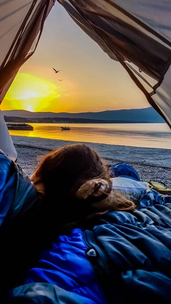 A girl lying in a tent set on the beach and watching the beautiful sunrise in Krk, Croatia. Sun is rising from behind the hill, turning sky orange. Calm surface of the sea reflects the sunbeams.