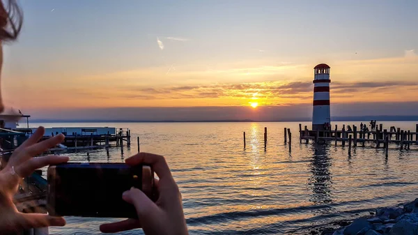 A young woman taking picture of a lighthouse located on the lake. Lighthouse has white and red stripes. Sun reflects itself in the calm water of the lake. Sunrise above the lake. Freedom and happiness